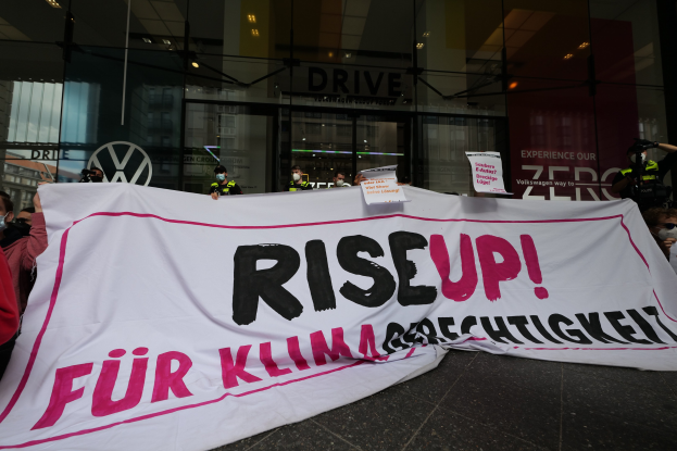 Eine Gruppe von Menschen mit Masken hält ein Transparent mit der Aufschrift "Steh Auf" und Schilder vor einem Glasfrontgebäude, wahrscheinlich protestieren sie gegen eine Entscheidung der deutschen Regierung bezüglich der Frauenrechte.