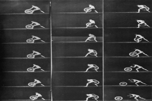 Ein Schwarz-Weiß-Foto eines Mannes im Rollstuhl, der ein Rad hält, ein weißes Hemd und dunkle Hosen trägt, mit entschlossener Miene und teilweise verdecktem Gesicht.