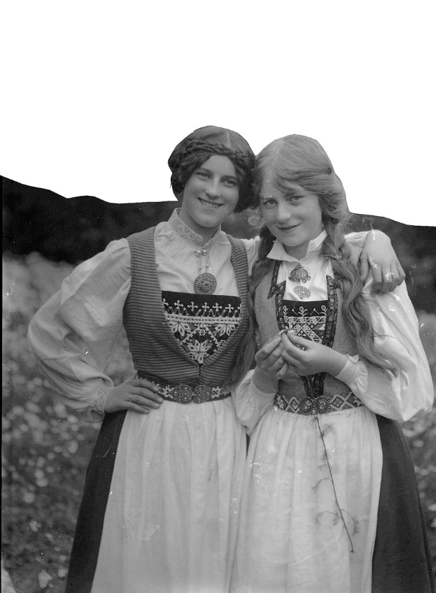 Zwei junge Frauen in traditionellen bayrischen Trachten mit weißer und schwarzer Stickerei und Perlen auf der Brust stehen lachend vor einem Hintergrund aus grünem Laub und einem hellen Himmel.