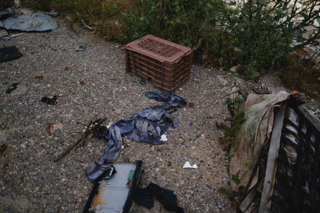 Haufen Müll, einschließlich eines Plastikbehälters und Kleidung, die auf dem Boden neben einem Zaun verstreut sind, mit Pflanzen und Gras im Hintergrund.