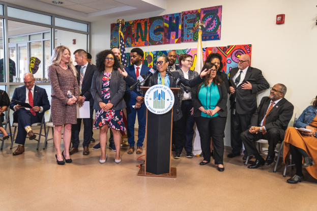 Gruppe von Menschen in einem Raum mit einem Podium, Mikrofon und einer Tafel mit der Aufschrift "Sancho Stadtrat tritt dem Stadtrat bei", umgeben von Stühlen, Flaggen und einem Feuerlöscher.