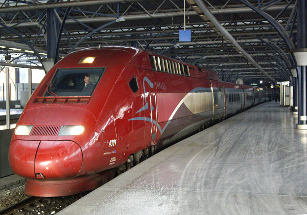 Ein roter Hochgeschwindigkeitszug steht auf einem Bahnsteig mit einer Plattform auf der rechten Seite, einige Menschen stehen und Säulen und Stangen im Hintergrund.