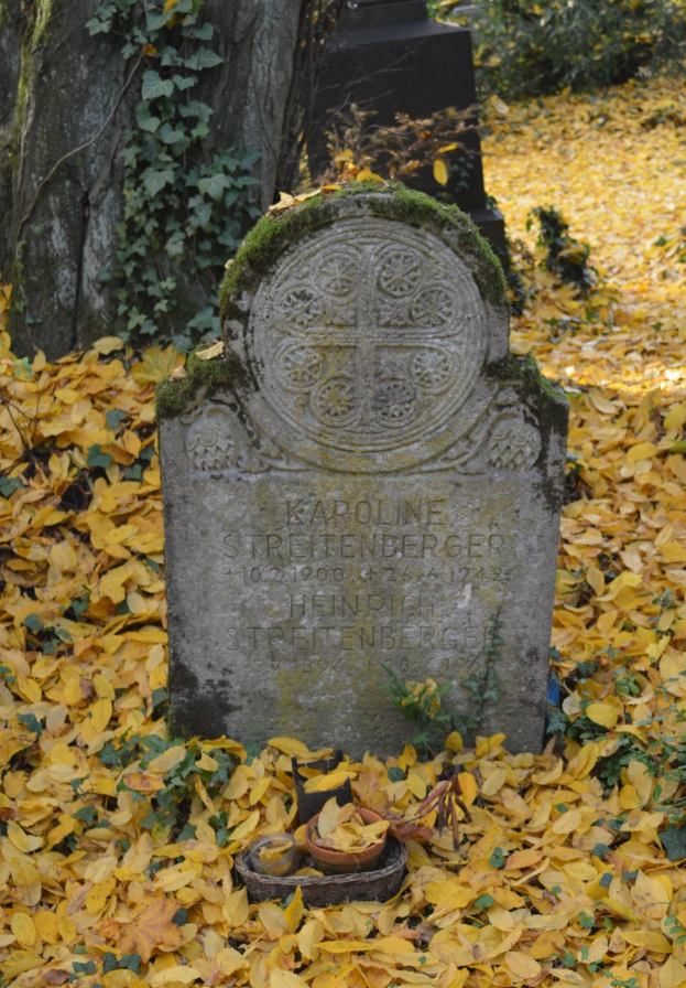 Ein Friedhof mit einem zentralen Grabstein, umgeben von gelben welken Blättern, Bäumen und Rankpflanzen im Hintergrund.