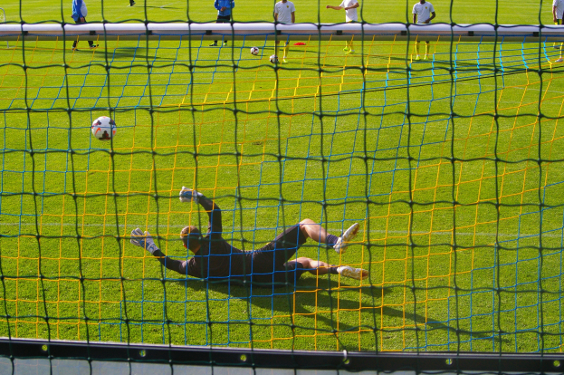 Ein Fußballspieler taucht ab, um vor einem Tor einen Ball zu halten, mit einem Netz im Vordergrund und einigen Menschen im Hintergrund, während mehrere Fußballschuhe auf dem Boden verstreut sind.