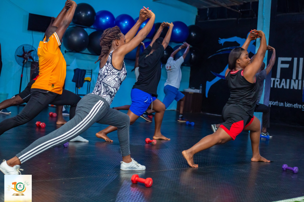 Gruppe von Menschen, die Yoga in einem Fitnessstudio mit Hanteln auf dem Boden machen, mit einem Banner "Fitness-Training in Ghana" im Hintergrund und diversen Gegenständen wie Luftballons und einem Ventilator.