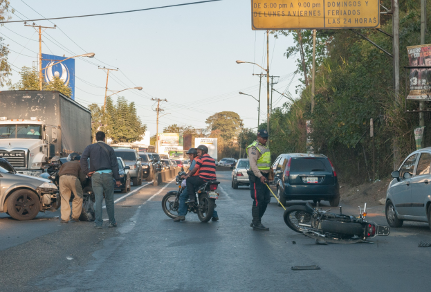 Eine Gruppe von Menschen steht neben einem verunglückten Motorrad am Straßenrand mit mehreren Fahrzeugen, darunter ein Lastwagen, im Hintergrund und Bäumen, Pfählen, Laternen, Schildern und Himmel.