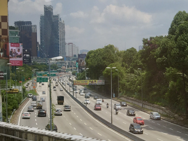 Eine vielbefahrene Autobahn mit zahlreichen Fahrzeugen, gesäumt von hohen Gebäuden, Straßenlaternen, Werbetafeln und Bäumen unter einem bewölkten Himmel.