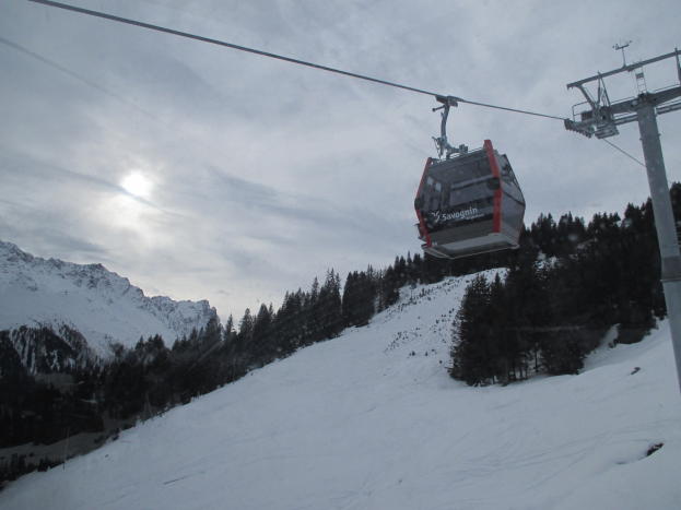 Eine Seilbahn fährt einen schneebedeckten Berg hinauf mit Bäumen im Hintergrund, unter einem bewölkten Himmel mit hellen Sonnenstrahlen.