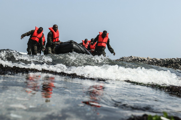 Gruppe von Menschen in Schwimmwesten auf einem Boot im Wasser, umgeben von Felsen auf der rechten Seite und einem klaren Himmel im Hintergrund, wahrscheinlich Teil eines Suchtrupps nach Uberlebenden.