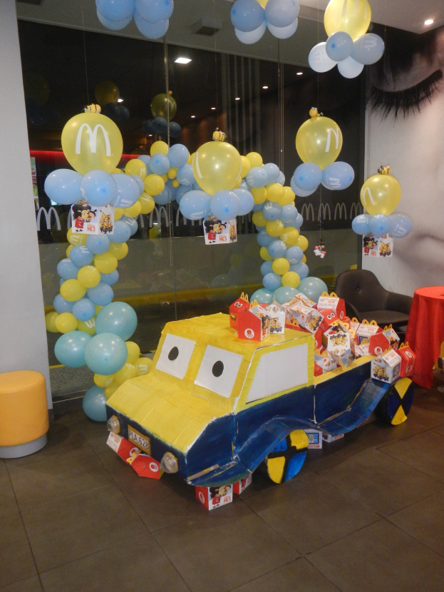 Eine McDonald's Geburtstagsparty mit einem gelben und blauen Lastwagen, der mit Ballons und anderen Dekoartikeln geschmückt ist, einem rot gedeckten Tisch mit Gegenständen, Hockern und einer Glaswand im Hintergrund, sowie einer Decke mit Lichtern, die eine festliche Atmosphäre schaffen.