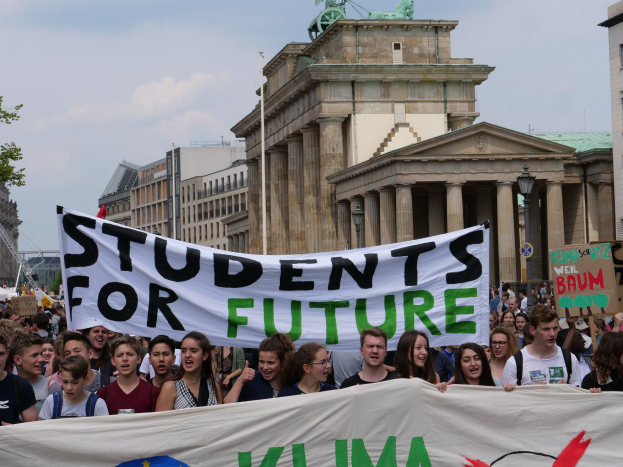 Eine Gruppe von Schülern marschiert in Berlin, die eine bunt bedruckte Fahne mit der Aufschrift "Students for Future" schwenken, mit Gebäuden, Bäumen und Himmel im Hintergrund.