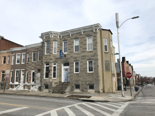Eine Häuserreihe an einer Straßenecke in einem neuen Wohnprojekt in Philadelphia, mit Fenstern, Türen, Treppen mit Geländern, Laternen, Schildern und Fahrzeugen, sowie Bäumen und einem bewölkten Himmel im Hintergrund.