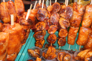Marktstand mit Spießen von Hähnchenflügeln und anderen Lebensmitteln in Plastikbehältern, mit Menschen und Flaschen im Hintergrund.