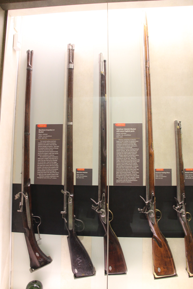 Vitrine mit verschiedenen Feuerwaffen, darunter ein Steinschlossgewehr, in einer Reihe mit Informationsschildern dahinter und einem unscharfen Objekt unten.