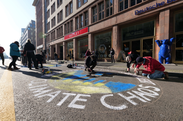 Eine Gruppe von Menschen sitzt vor einem Gebäude mit Fenstern und Namensschildern auf dem Boden, umgeben von Flaschen und anderen Gegenständen, mit Bäumen und einem klaren blauen Himmel im Hintergrund, bei einer Klimawandel-Demonstration in Berlin.