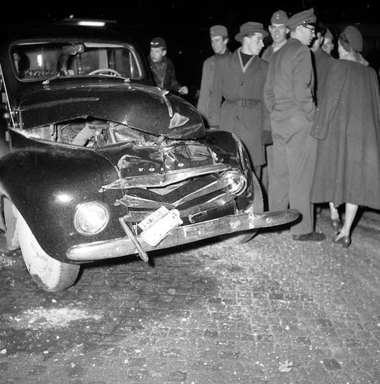 Schwarzes und weißes Bild eines schwer beschädigten Autos am Straßenrand mit einer Gruppe von Menschen, die es inspizieren.