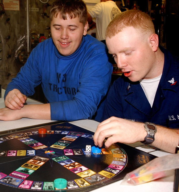 Zwei Männer sitzen an einem Tisch und spielen ein Brettspiel mit Würfeln und anderen Gegenständen auf dem Tisch, während im Hintergrund ein paar Menschen stehen.
