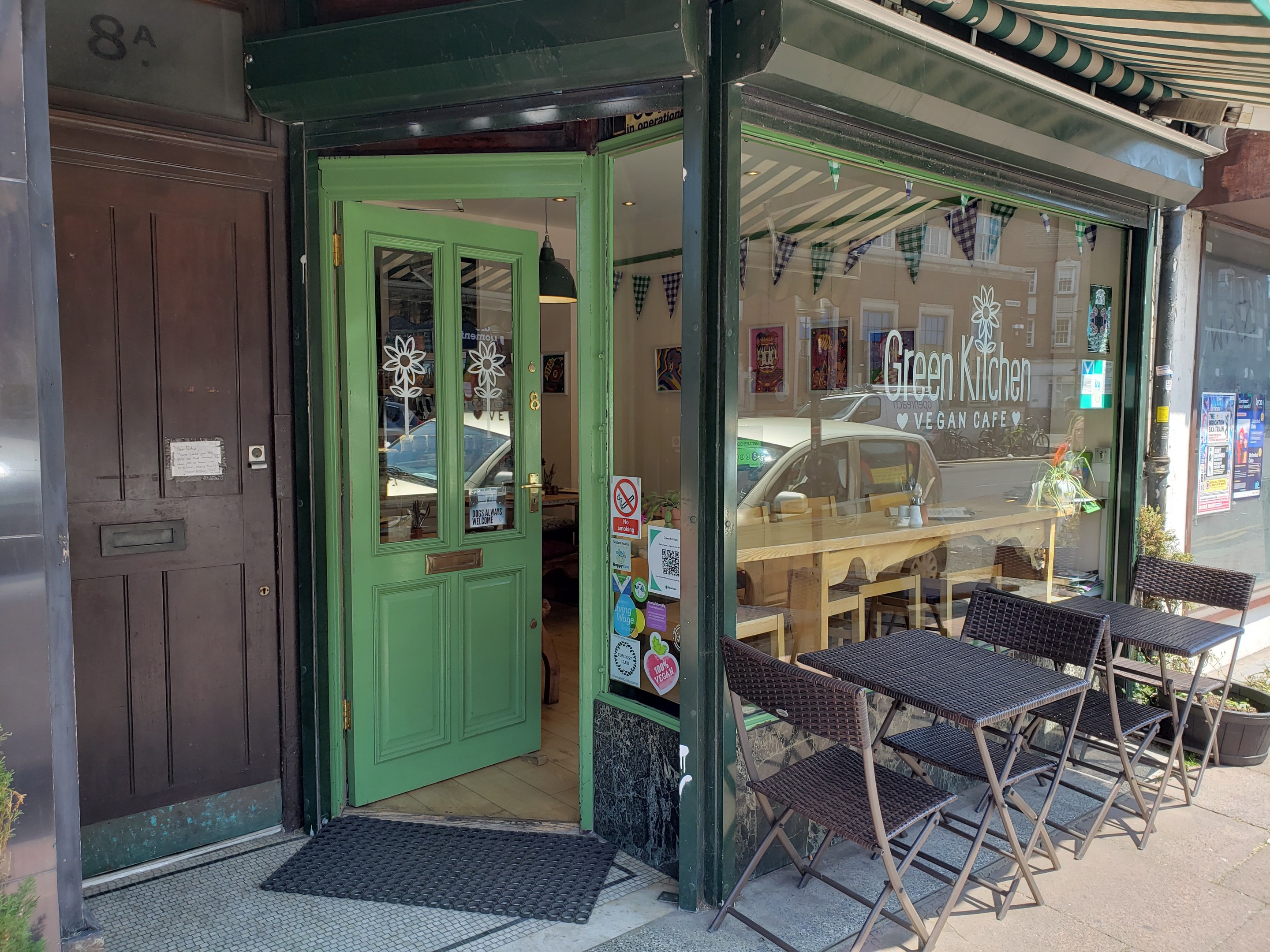 Außensitzbereich des Green Kitchen Vegan Cafe mit Tischen, Stühlen, Pflanzen, einer Fußmatte und einer Glastür mit Postern, der in ein Inneres mit Tischen, Stühlen und einer Hängelampe führt.