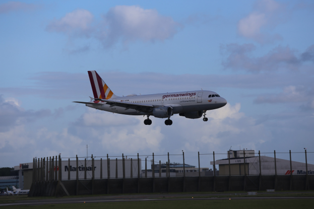 Germanwings Airbus A320-200 beim Start am Frankfurter Flughafen mit Gras darunter, einem Zaun im Vordergrund, Gebäuden und Bäumen im Hintergrund und einer bewölkten Himmel über dem Flugzeug.