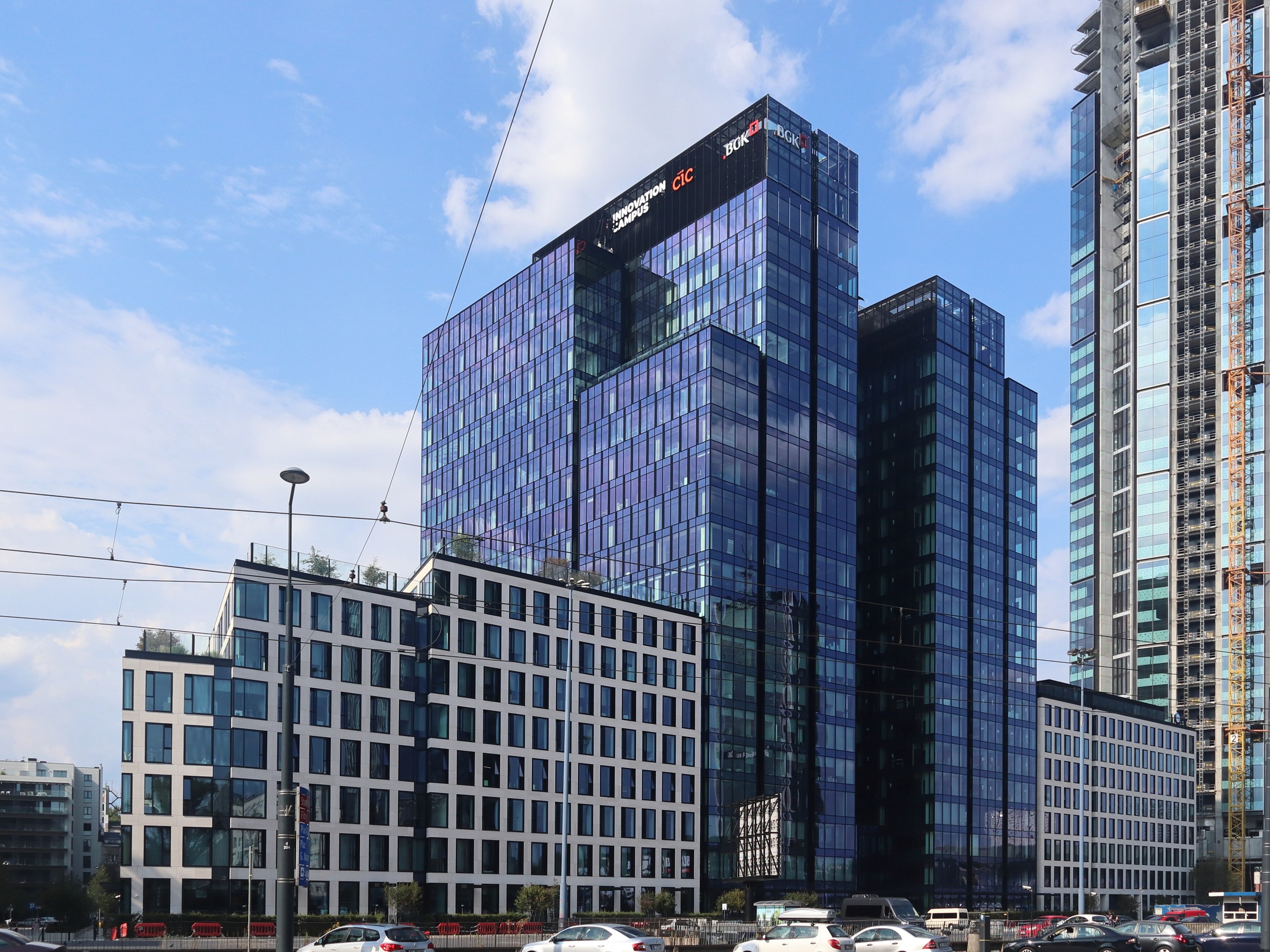 Außenansicht des Deutschen Bank-Hauptsitzes in Frankfurt, Deutschland, mit einem mehrstöckigen Gebäude mit zahlreichen Fenstern, umgeben von städtischer Infrastruktur wie Straßenlaternen, Fahrzeugen und Grünflächen unter einem bewölkten Himmel.