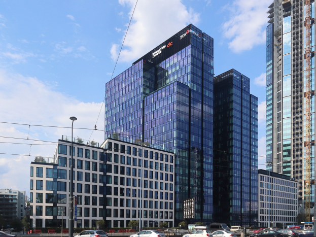 Außenansicht des Deutschen Bank-Hauptsitzes in Frankfurt, Deutschland, mit einem mehrstöckigen Gebäude mit zahlreichen Fenstern, umgeben von städtischer Infrastruktur wie Straßenlaternen, Fahrzeugen und Grünflächen unter einem bewölkten Himmel.