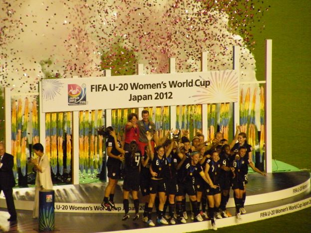 Eine Gruppe von Frauenfußballspielerinnen in schwarzen Uniformen feiert ihren Sieg auf einer Bühne, umgeben von einer Menge mit Kameras, mit einem dekorierten Hintergrund und einem grasbewachsenen Boden.