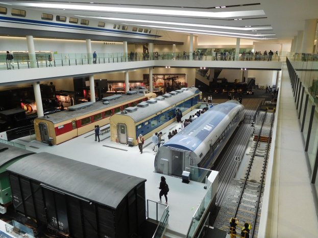 Ein großer Museumsraum mit verschiedenen Bahnen in unterschiedlichen Größen und Farben, mit Menschen, die die Ausstellungsstücke und architektonische Elemente wie Geländer, Säulen und Deckenleuchten betrachten.