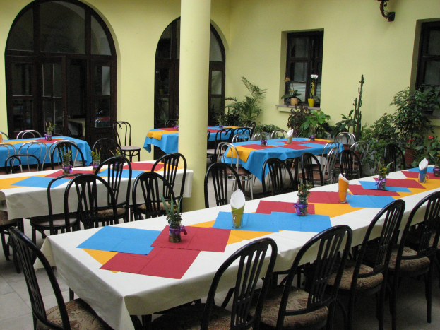 Eine Kantine mit Tischen und Stühlen, die für eine Mahlzeit gedeckt sind, geschmückt mit bunten Tischdecken und Blumenvasen, mit einer Wand aus Fenstern und Pflanzen im Hintergrund.