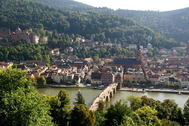 Eine malerische Aussicht auf Heidelberg, Deutschland, mit einer Brücke über einen Fluss, Fahrzeugen auf der Straße, Gebäuden mit Fenstern, Bäumen, Hügeln und einem klaren blauen Himmel.