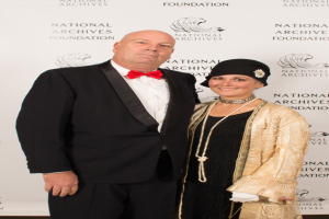 Ein Mann in einem schwarzen Anzug mit einem weissen Hemd und schwarzer Fliege steht neben einer Frau in einem eleganten Kleid mit einem gemusterten Schal auf einem roten Teppich, mit einem weissen Banner mit Text im Hintergrund.