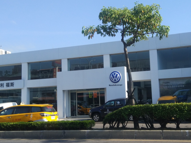 Ein Volkswagen-Händler mit Glaswänden und -fenstern, der Autos zeigt, die davor geparkt sind, umgeben von Pflanzen und einem Baum unter einem sichtbaren Himmel.