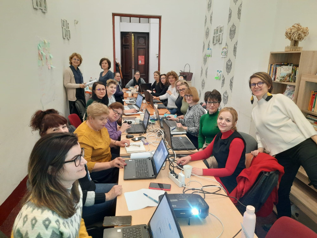 Eine Gruppe von Frauen in einem Workshop-Setting mit Laptops, Papieren und anderen Gegenständen auf einem Tisch, umgeben von Stühlen mit Jacken, einem Schrank mit Büchern und einer Vase und Papieren an der Wand.