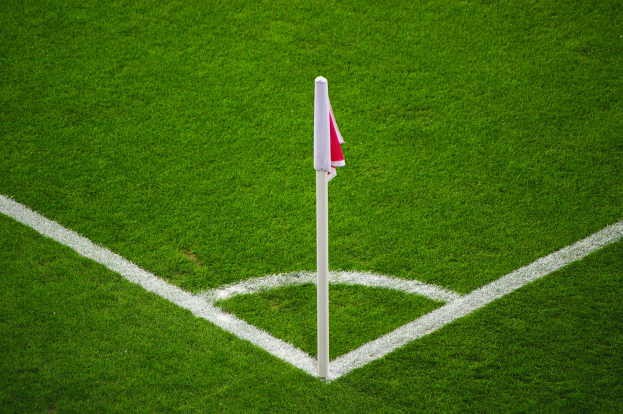 Ein Fußballfeld mit grünem Rasen, weißen Begrenzungslinien und einem weißen Pfosten mit einer roten und weißen Flagge in der Mitte des Strafraums.