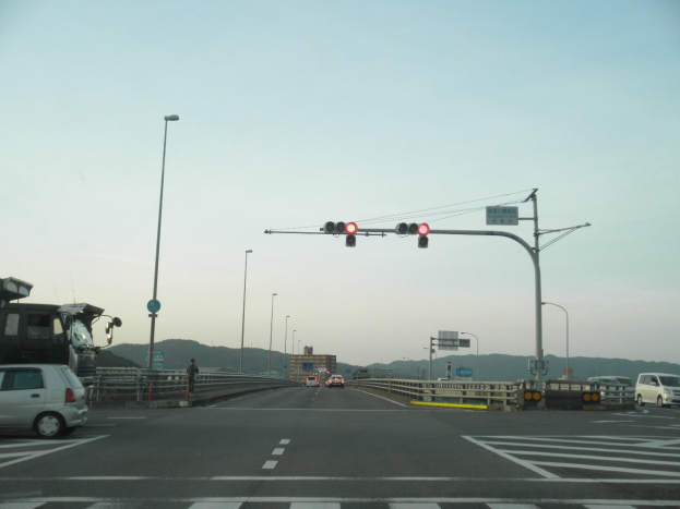 Ein Auto fährt auf einer Autobahn an einer Kreuzung mit einer Ampel vorbei, umgeben von Fahrzeugen, einem Fußgänger auf einem Gehweg, Geländern, Laternen, Verkehrszeichen, Straßenschildern, Gebäuden, Hügeln und einem klaren blauen Himmel.