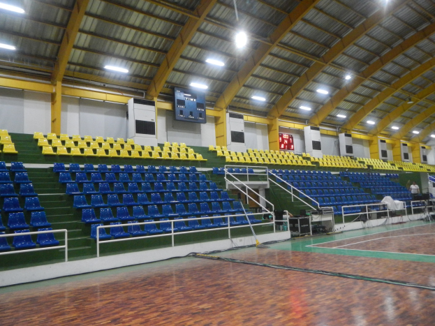 Große Sporthalle mit Holzboden, blauen und gelben Sitzplätzen, Geländern, Treppen mit Geländern, wandmontierten Bildschirmen, Deckenleuchten und einigen Anwesenden.
