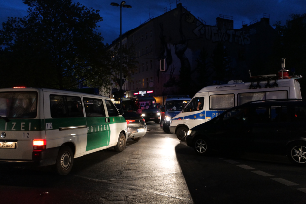 Eine Gruppe von Polizeiwagen, darunter ein Van, ein Auto und ein Rad, geparkt an der Seite einer Straße bei Nacht mit Gebäuden, Bäumen, Laternen und einem sternenklaren Himmel im Hintergrund.