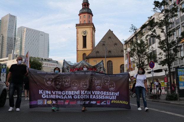 Eine Gruppe maskierter Personen marschiert mit einem Transparent die Straße entlang, mit einem geparkten Auto auf der linken Seite, Gebäuden, Bäumen, Schildern, Pfählen und einem Uhrenturm im Hintergrund unter einem klaren blauen Himmel.