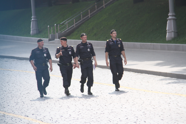 Drei Polizisten in Uniform und mit Mützen, die eine Straße mit Geländern, Treppen und Gras im Hintergrund entlanggehen.