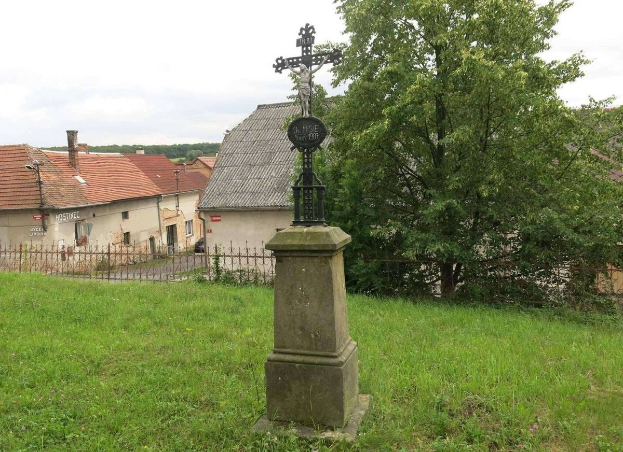 Ein großes Kreuz steht als Holocaust-Mahnmal in einem eingezäunten Grasfeld, mit Bäumen, Häusern und einem bewölkten Himmel im Hintergrund.