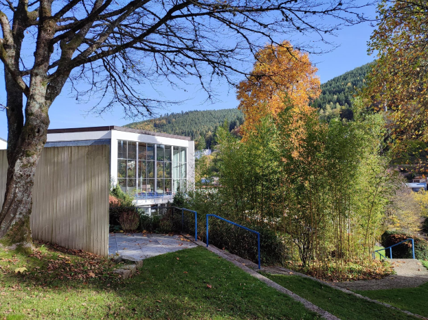 Außenansicht des Universitätsgebäudes Stuttgart mit einem Baum davor, umgeben von Gras, trockenen Blättern, Pflanzen und einem Geländer, mit Hügeln und einem klaren blauen Himmel im Hintergrund.
