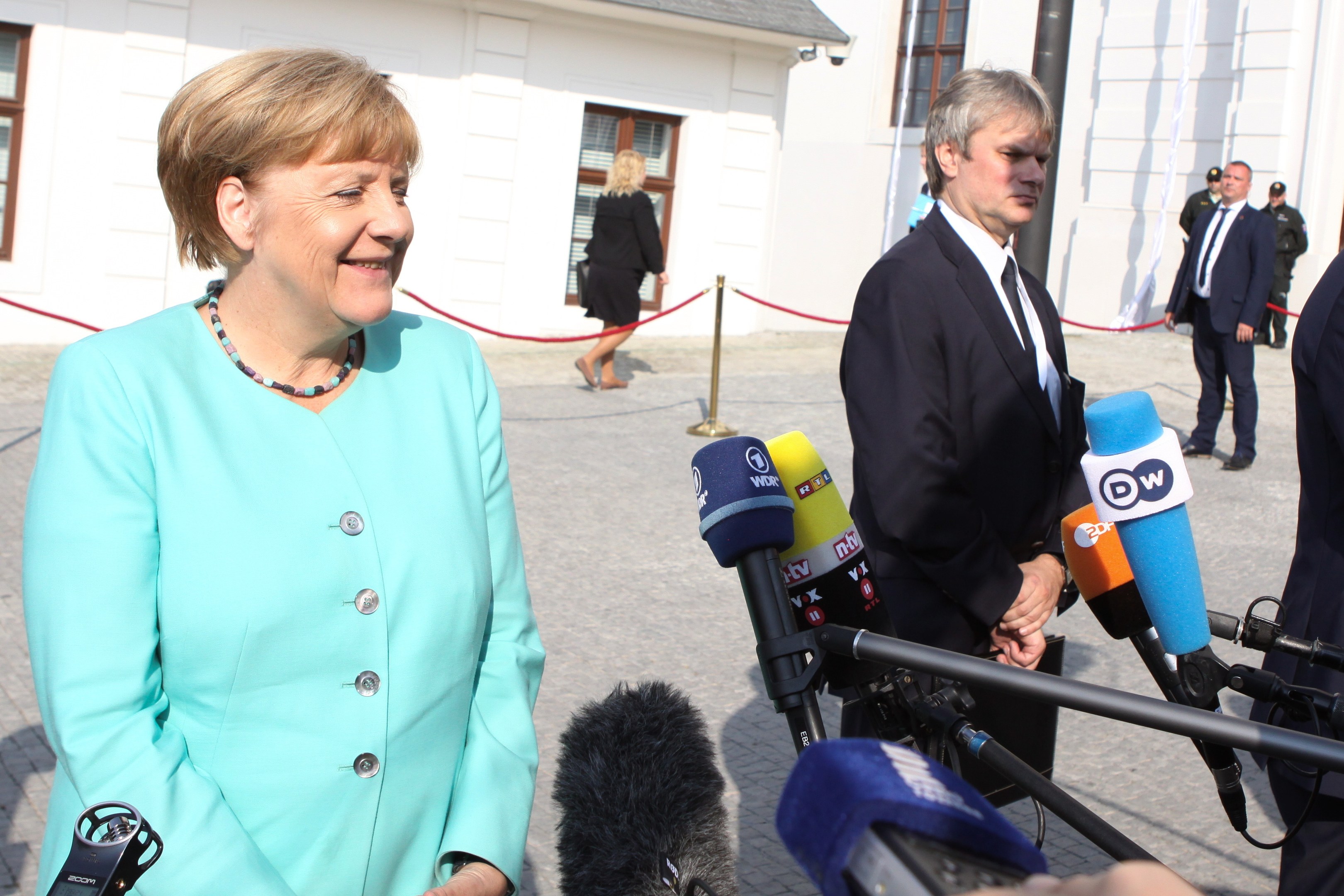 Bundeskanzlerin Angela Merkel beim Sprechen mit Reportern im Freien, umgeben von einer Gruppe mit Mikrofonen und einer Absperrung, unter einem bewölkten Himmel.