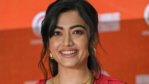 Eine Frau in einem roten Kleid mit einem eleganten Dutt und dezentem Make-up steht lächelnd da, trägt schwarze High Heels und hat ein hellgelbes Banner mit der Aufschrift "Sonam Kapoors Beste Schönheitsblicke vom Cannes Film Festival" im Hintergrund.