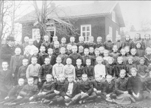 Schwarzes und weißes Foto einer Gruppe von Kindern, die vor einem Haus mit Fenstern posieren, umgeben von Bäumen und einem klaren Himmel, mit Text unten, der "1930er-Schulkinder" lautet.