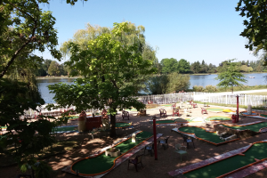 Minigolf-Platz in einem Park mit grünem Gras, Bäumen und einem Zaun, umgeben von Stühlen und Tischen, mit einem Gewässer und einem Boot im Hintergrund unter dem Himmel.