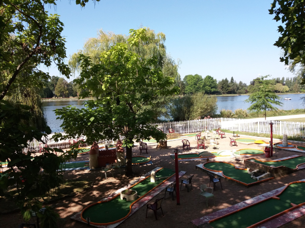 Minigolf-Platz in einem Park mit grünem Gras, Bäumen und einem Zaun, umgeben von Stühlen und Tischen, mit einem Gewässer und einem Boot im Hintergrund unter dem Himmel.