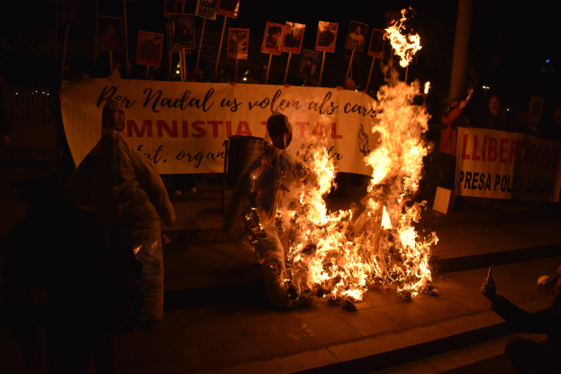 Eine Gruppe von Menschen steht um ein hell brennendes Lagerfeuer auf der Straße in der Nacht, mit Schildern im Hintergrund und einem Müllcontainer im Vordergrund.