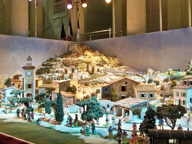 Eine detaillierte Museumsausstellung einer Krippe mit Figuren, Gebäuden, Bäumen, einem Kirchturm, Flaggen und beleuchteten Elementen.