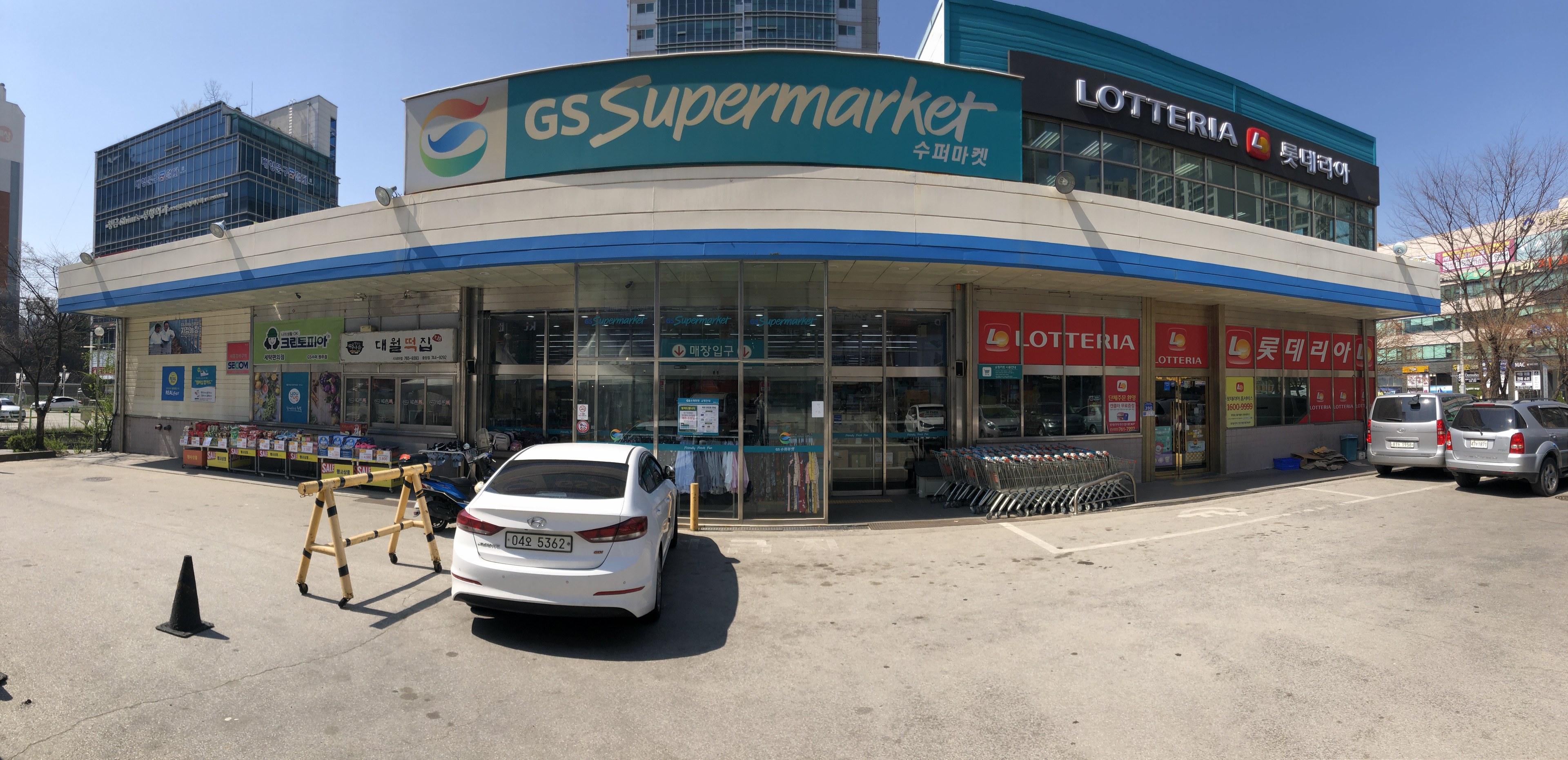 Ein Auto steht vor einem "GS Supermarkt" mit anderen Fahrzeugen, Verkehrskegeln und verstreuten Gegenständen auf der Straße, vor Häusern, Bäumen und einem klaren blauen Himmel.
