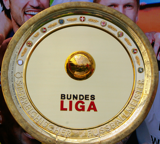 Ein Mann hält ein goldenes Schild mit 'Bundesliga' hoch, im Hintergrund eine Fahne mit drei Personen.