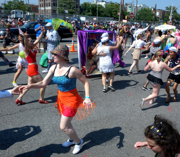 Eine Gruppe von Menschen, die eine Stra√e entlangmarschieren, einige mit Mötzen und Schirmen, andere tanzend, mit einer Fahne, Verkehrskegeln, Fahrzeugen, Laternenmästen, Schildern, Bäumen, Gebäuden und einem klaren blauen Himmel im Hintergrund.